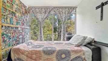 Un dormitorio con una cama, una estantería, una ventana con cortinas y una cruz en la pared.