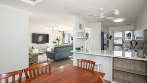 Ett modernt kök med en köksö i mitten, en matplats med ett träbord och stolar, ett vardagsrum med en grå soffa och en platt-TV samt en takfläkt.