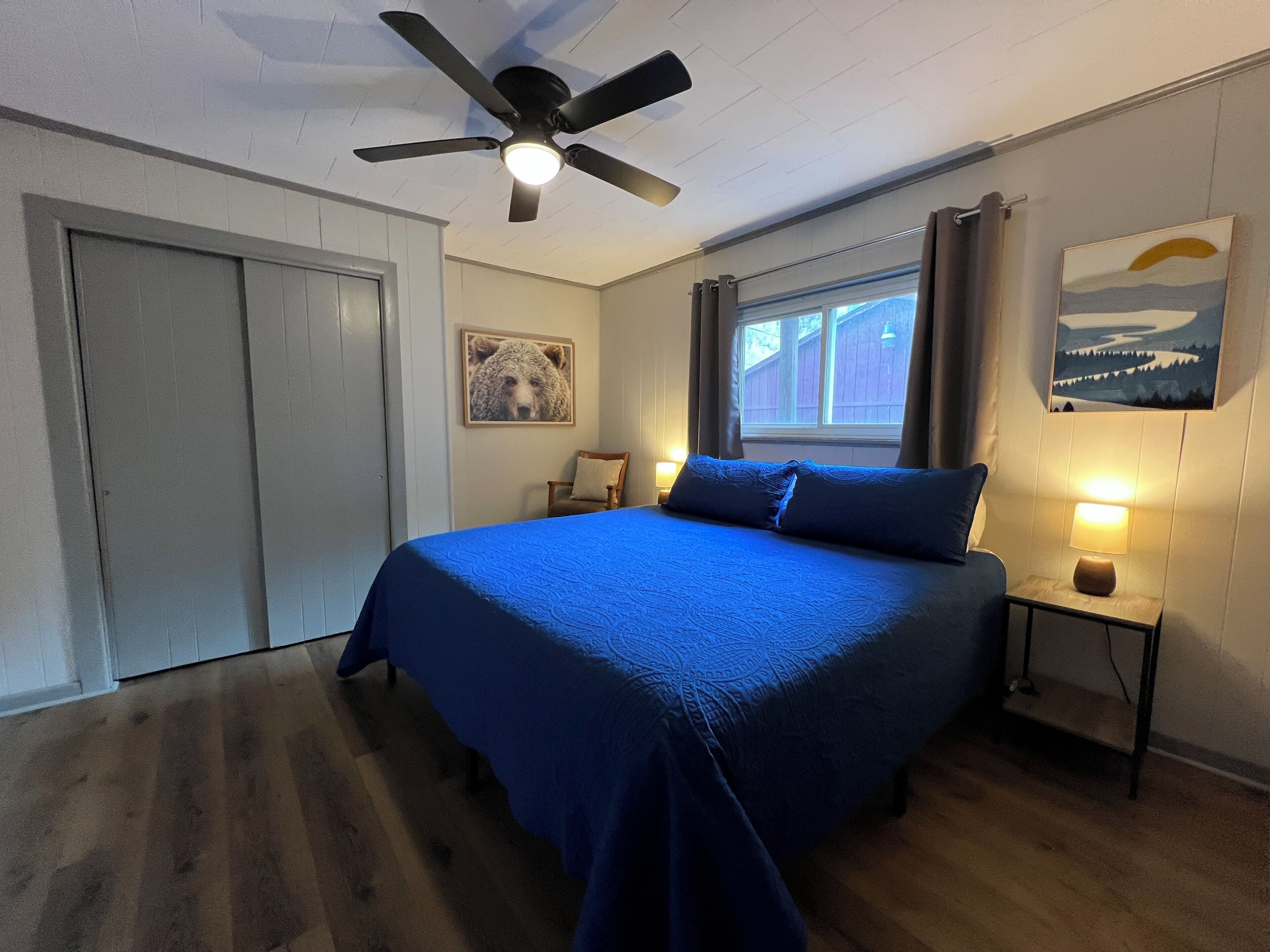 A bedroom with a bed, a ceiling fan, a window with curtains, a bedside lamp, and a wall-mounted picture.
