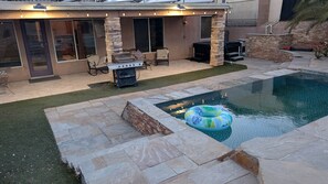 En trädgård med en pool, en stenbelagd uteplats, en utomhusgrill och utemöbler.