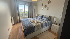 Una camera da letto con un letto grande, un comodino, una lampada, una finestra con tende e vista sugli edifici esterni.