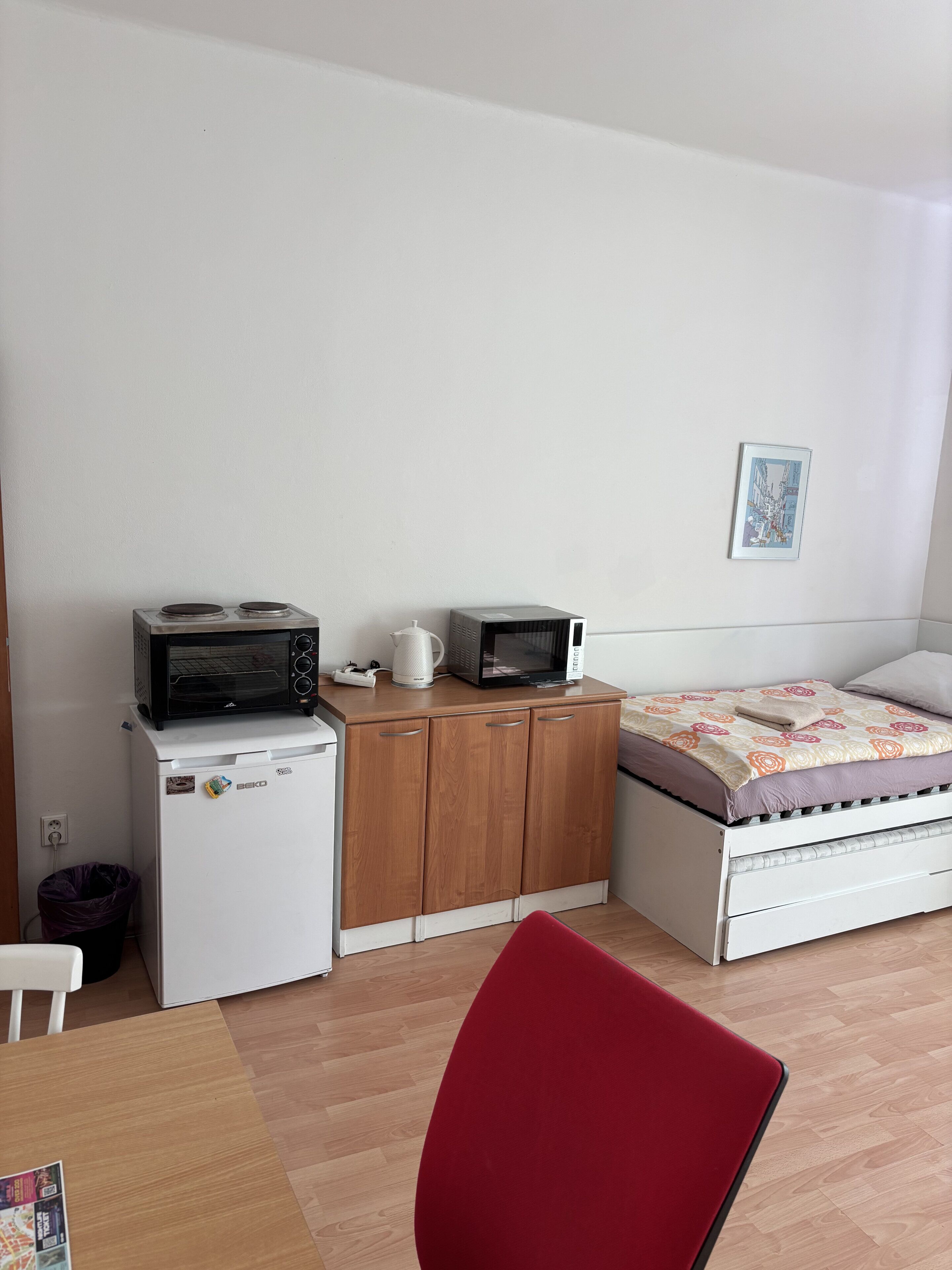 A small, compact room with a bed, a kitchenette with a microwave and a small fridge, a wooden cabinet, and a red chair.