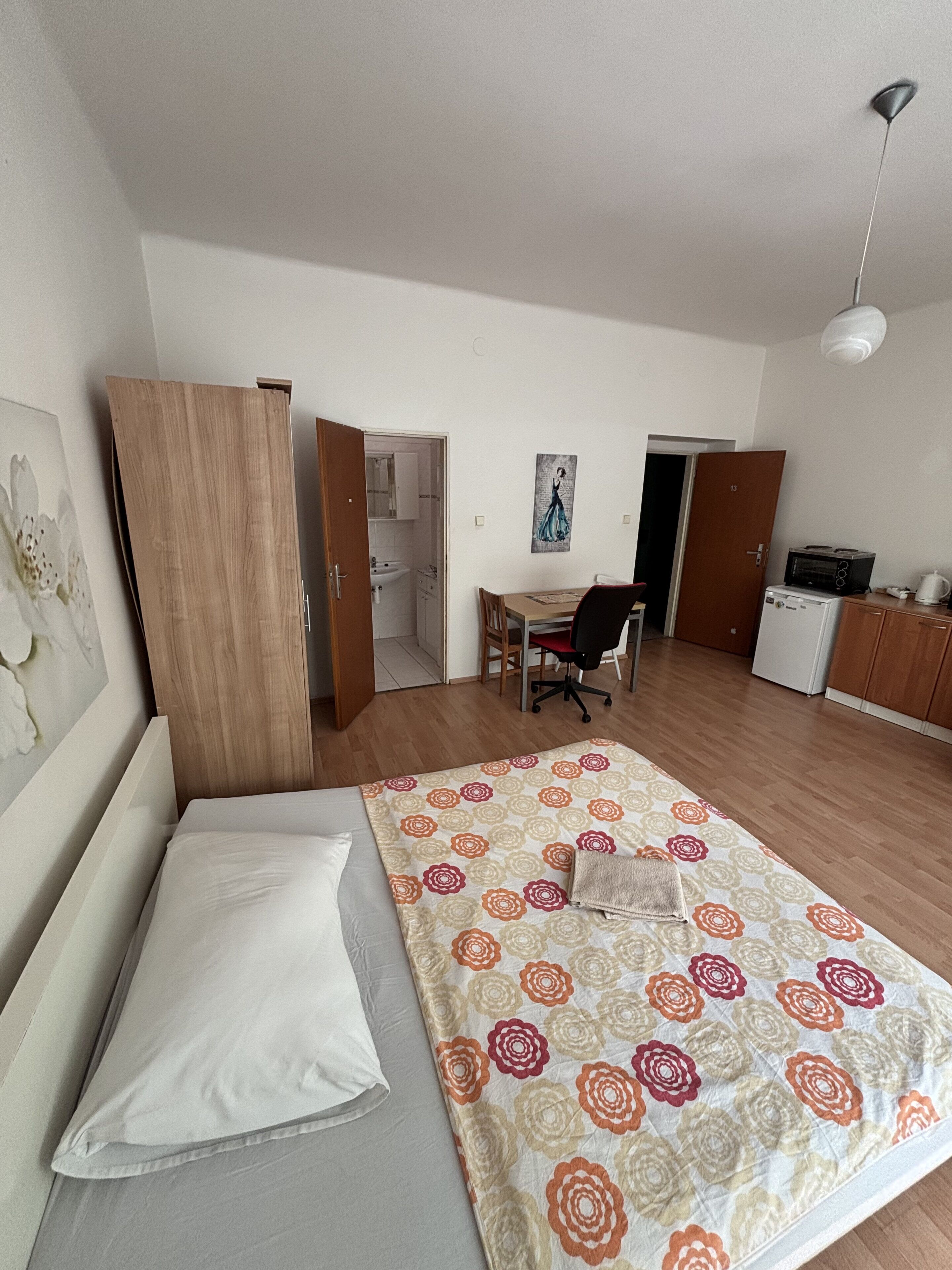 A bedroom with a bed, a desk, a chair, and a kitchenette.