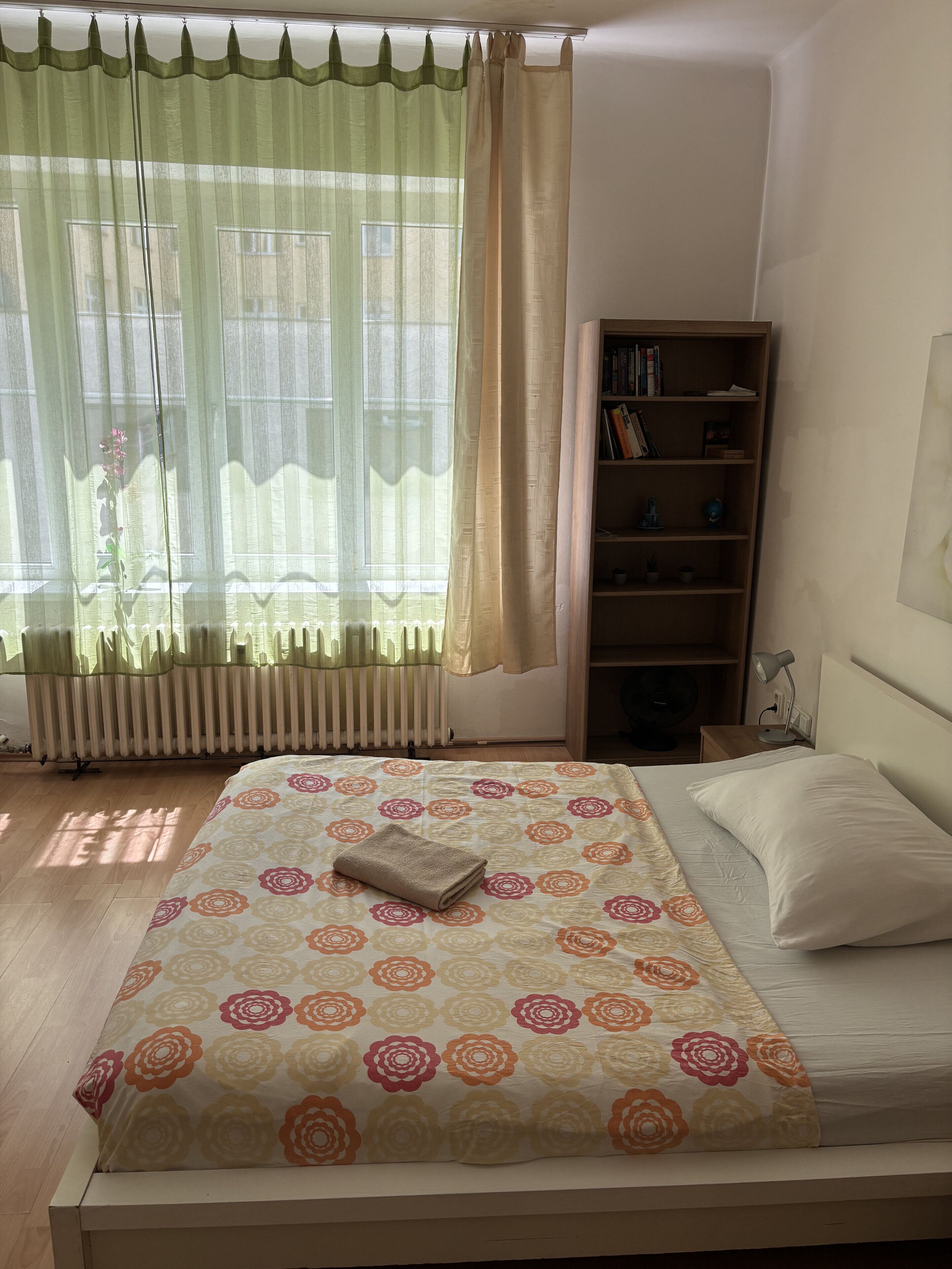 Ein Schlafzimmer mit einem Bett, einem Heizkörper, einem Bücherregal und einem Fenster mit Vorhängen.