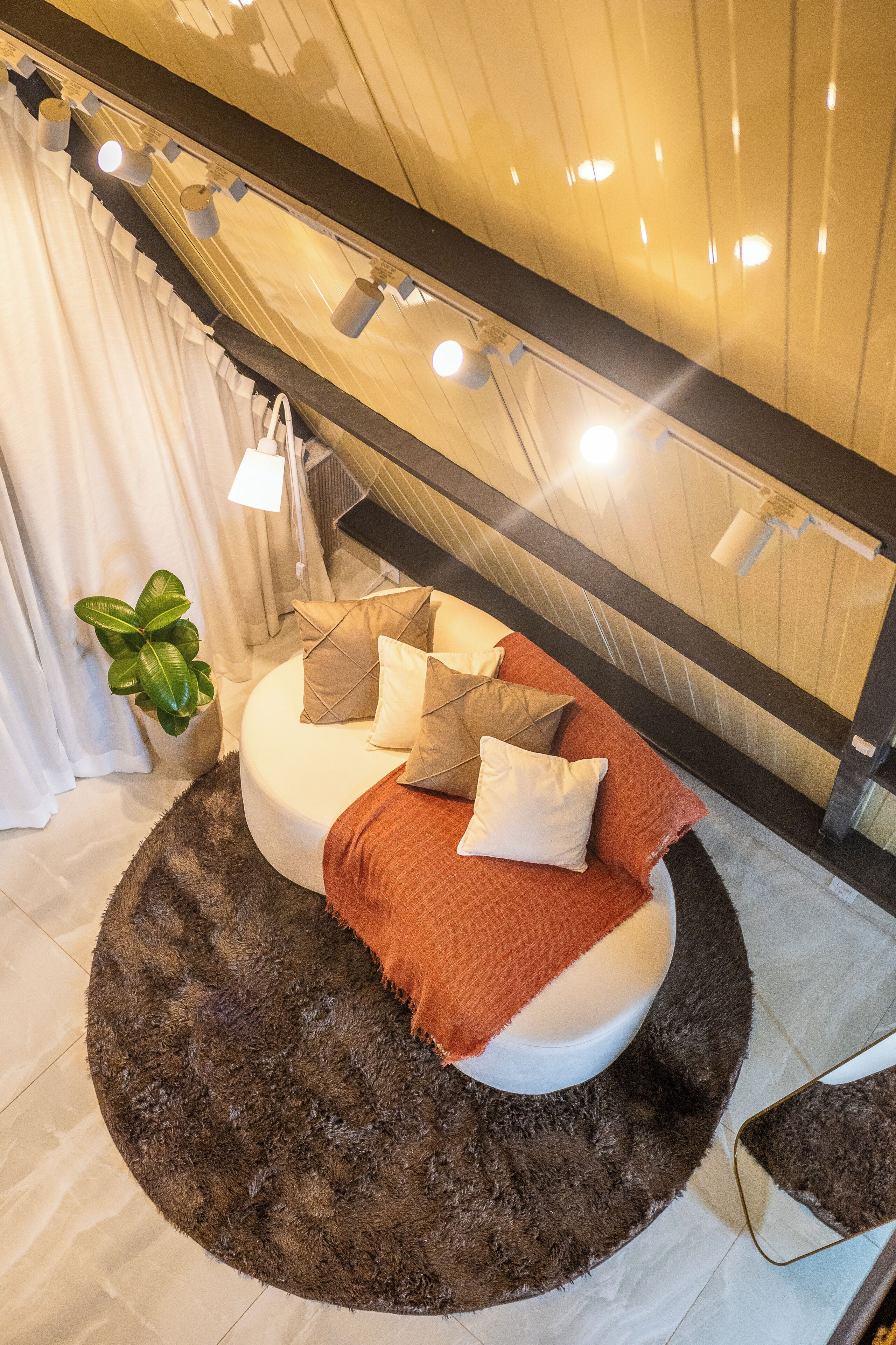 Quarto moderno com cama redonda, várias almofadas, tapete escuro, planta em vaso e janela com cortinas.