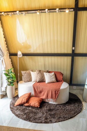A modern living room with a round white sofa, multiple cushions, a dark shag rug, a floor lamp, and a potted plant.