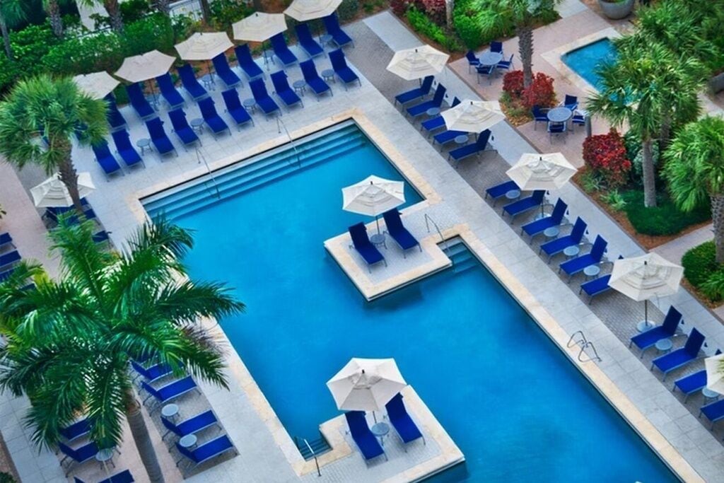 A pool area with multiple lounge chairs and umbrellas, surrounded by palm trees.