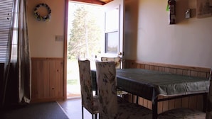 A room with a wooden ceiling, a table with a green tablecloth, two floral-patterned chairs, a door leading outside, and a wall-mounted fire extinguisher.