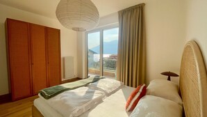 A bedroom with a bed, a wooden wardrobe, a radiator, a window with curtains, and a view of mountains. - 2-bedroom apartment in tranquil Brenzone sul Garda with AC (Brenzone sul Garda)