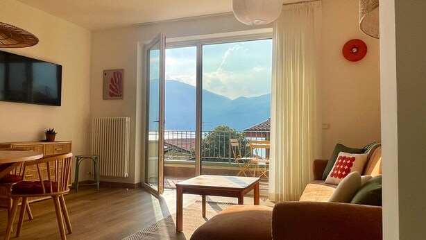 A living room with a dining area, a sofa, a TV, and a view of mountains through large windows. - 2-bedroom apartment in tranquil Brenzone sul Garda with AC (Brenzone sul Garda)