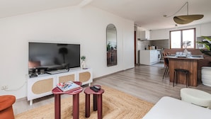 A modern living room with a flat-screen TV, a dining area with a wooden table, and a kitchen in the background. - Le Grandval - Spacious 3 rooms (Reims)