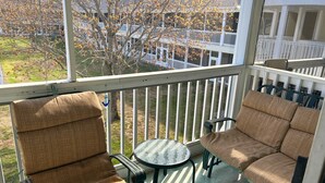 Ett takförsett utomhusområde med sittplatser, bestående av två stolar och ett litet bord, med utsikt över ett bostadsområde med träd och ett vitt staket.