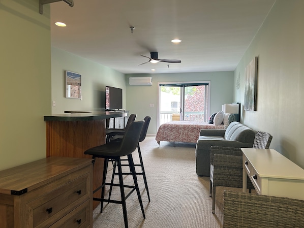 A room with a bed, a desk, chairs, a television, and a ceiling fan.