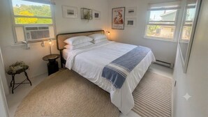 Een slaapkamer met een bed, nachtlampjes, een klein krukje, een radiator, en een raam met uitzicht op groen.