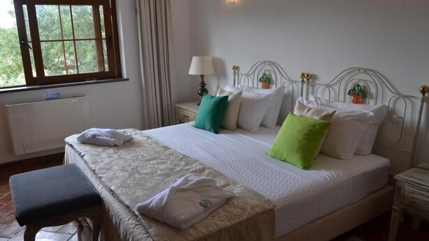 Una cama bien tendida con una colcha estampada, una mesita de noche con una lámpara y una ventana con cortinas.