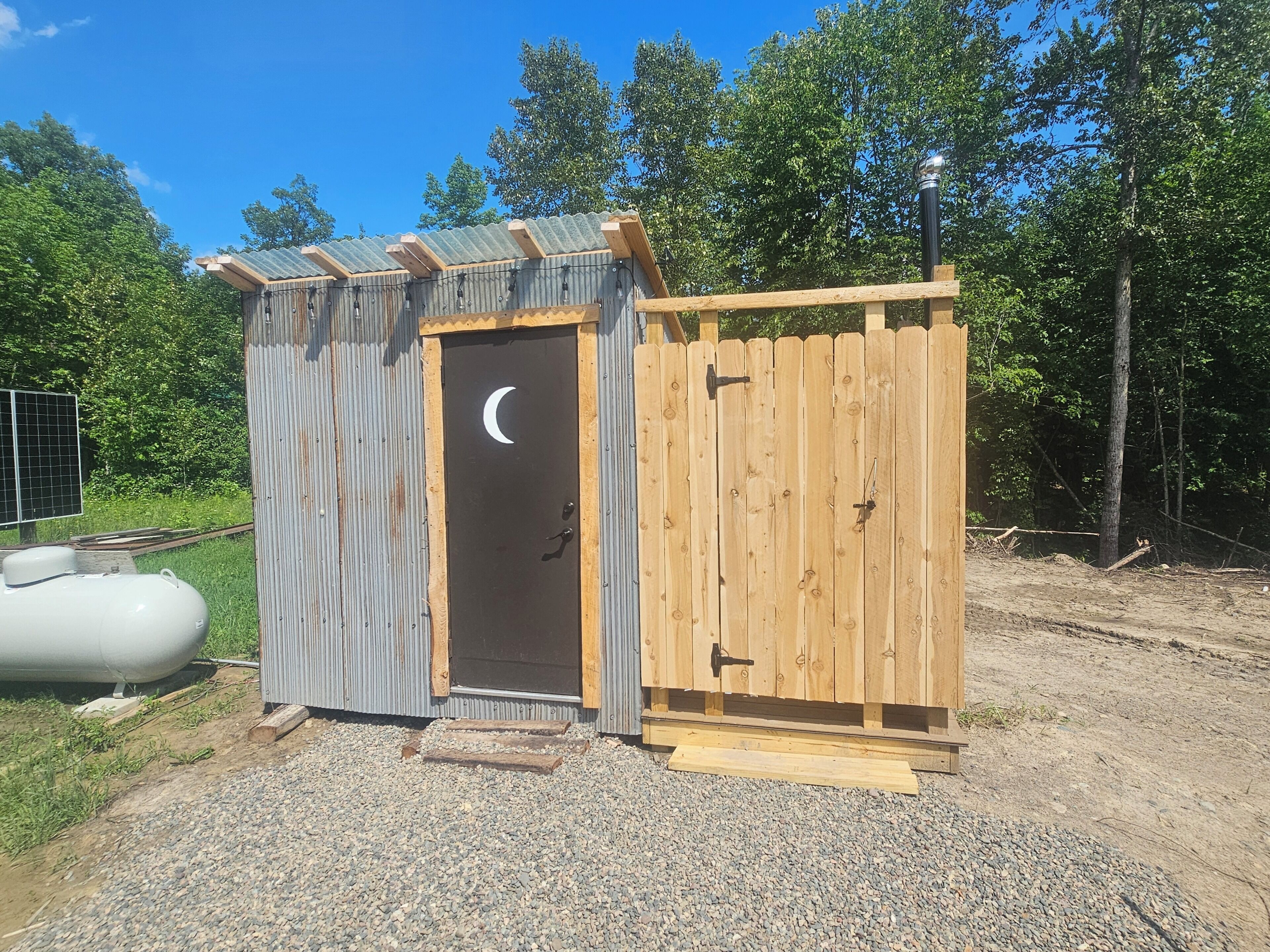 A small wooden structure with a door featuring a crescent moon design, a wooden fence, and a gravel ground.