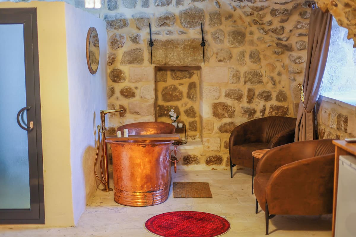 Quarto com paredes de pedra, uma banheira de cobre grande, uma poltrona de couro marrom e uma mesa de madeira.