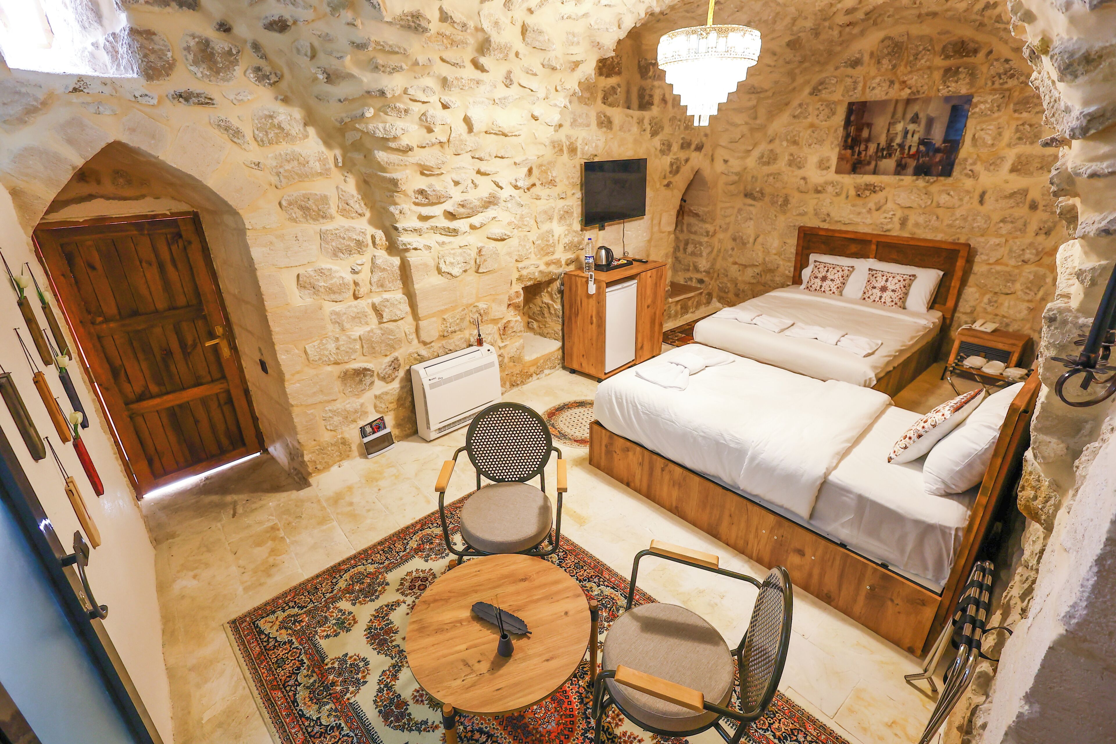 A stone-walled bedroom with a bed, two chairs, a small table, a TV, and a radiator.