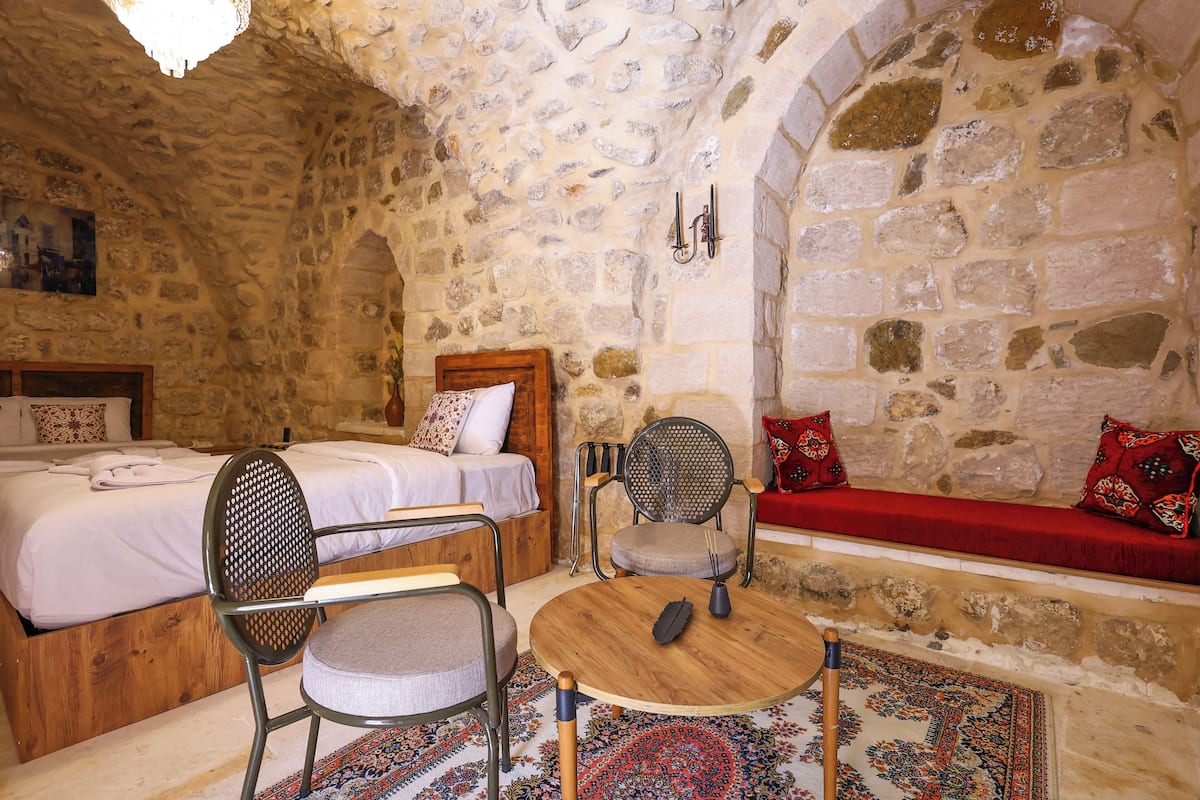 Quarto com paredes de pedra, uma cama, uma mesa de madeira, uma cadeira e um banco.