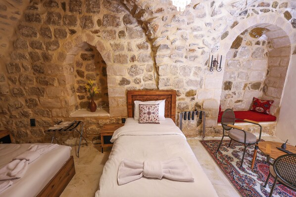 Una habitación con paredes de piedra, una cama, una silla, una mesita pequeña y un banco.