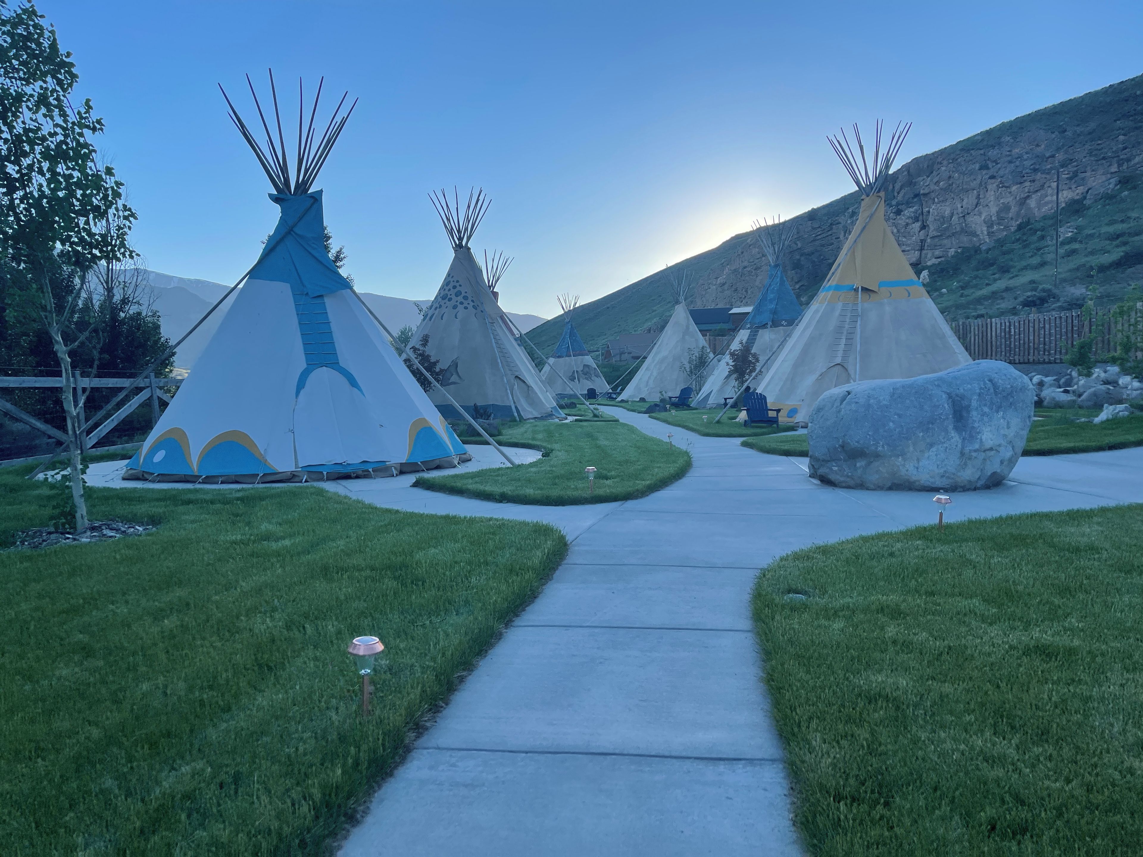 Un sendero conduce a varias tiendas tipi, con un área de césped y una montaña al fondo.
