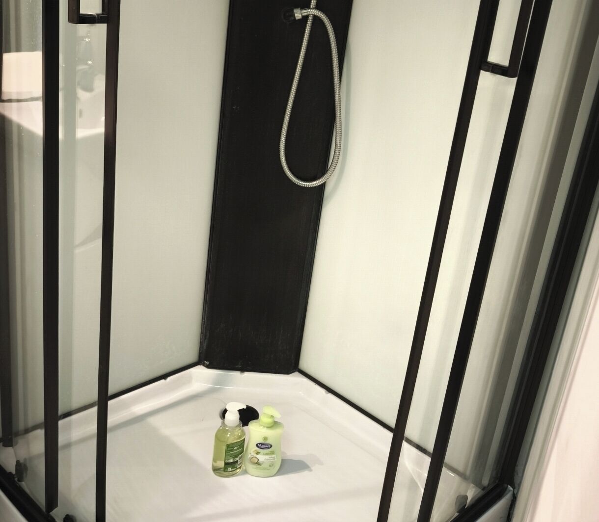 A corner shower with a handheld showerhead, a white floor, and two bottles of shampoo and conditioner.