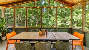 En overdækket terrasse med et træbord og orange stole, omgivet af store vinduer med udsigt over en skov.