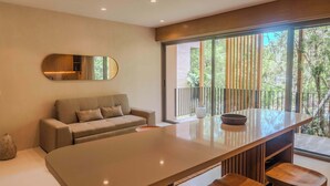 A modern interior with a sofa, a large mirror, a dining area with a table and stools, and a balcony with a view of greenery. - Luxury 1BR Aflora Retreat in Aldea Zama with  pool (Tulum)