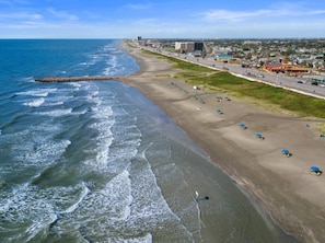 Ein Küstenstrand mit einem in das Meer führenden Steg, einer parallel zum Strand verlaufenden Straße und Gebäuden im Hintergrund.