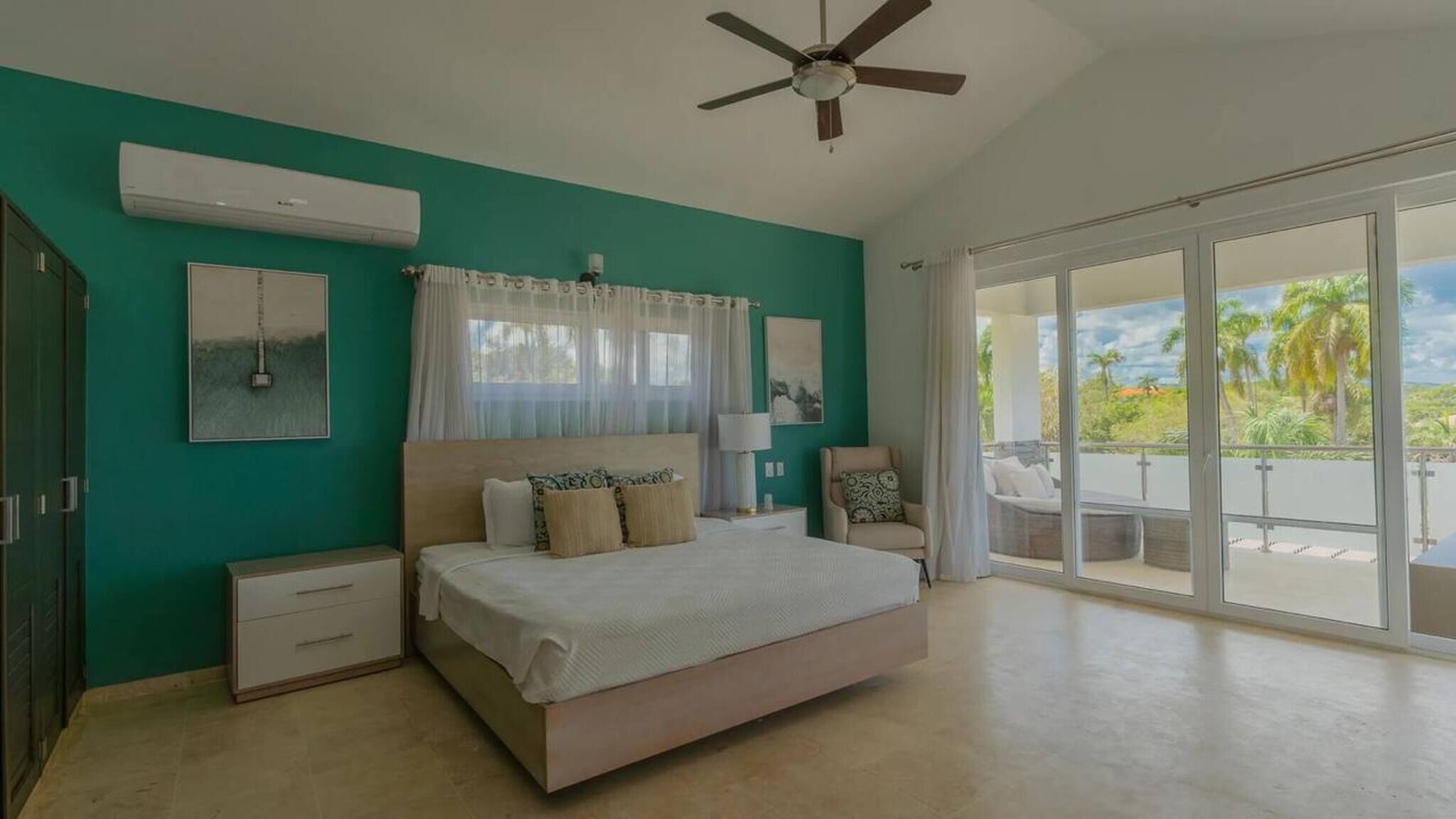 Um quarto com cama, ventilador de teto, janela com cortinas, mesa de cabeceira, cadeira e vista para o exterior.