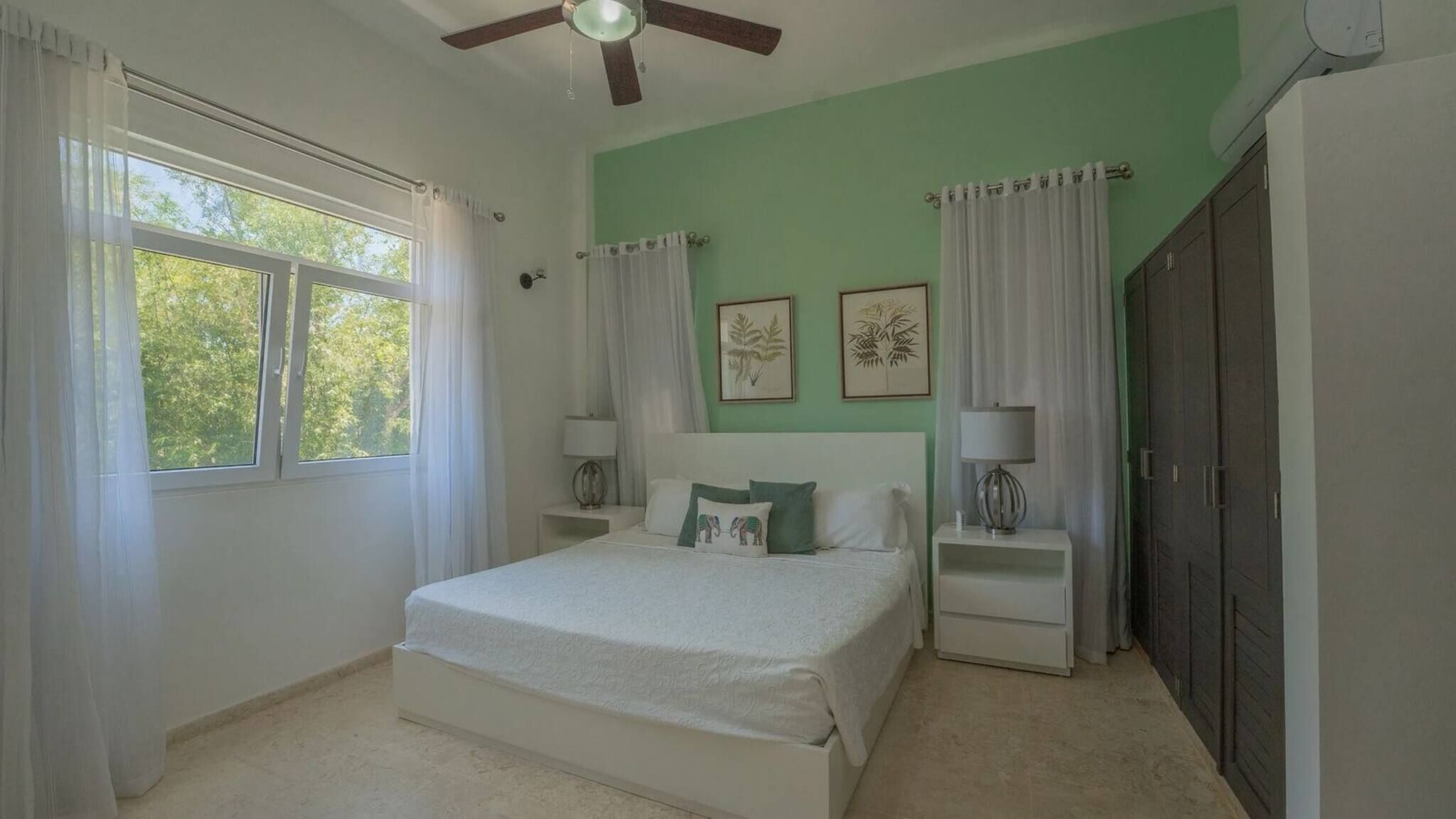 A bedroom with a bed, two bedside tables, a ceiling fan, a window with curtains, and a wardrobe.