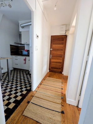 A narrow hallway with a checkered floor, a wooden door, and a kitchen area with a microwave. - right by the sea (Saint-Valery-en-Caux)