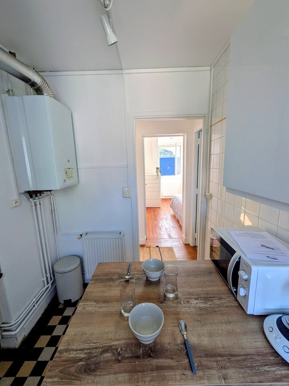 Een keuken met een houten tafel, witte apparaten en zicht op een slaapkamer.