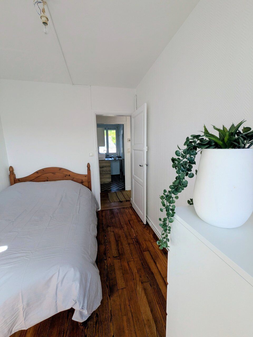 Um quarto com cama de madeira, parede branca, planta em vaso e porta que dá para uma cozinha.