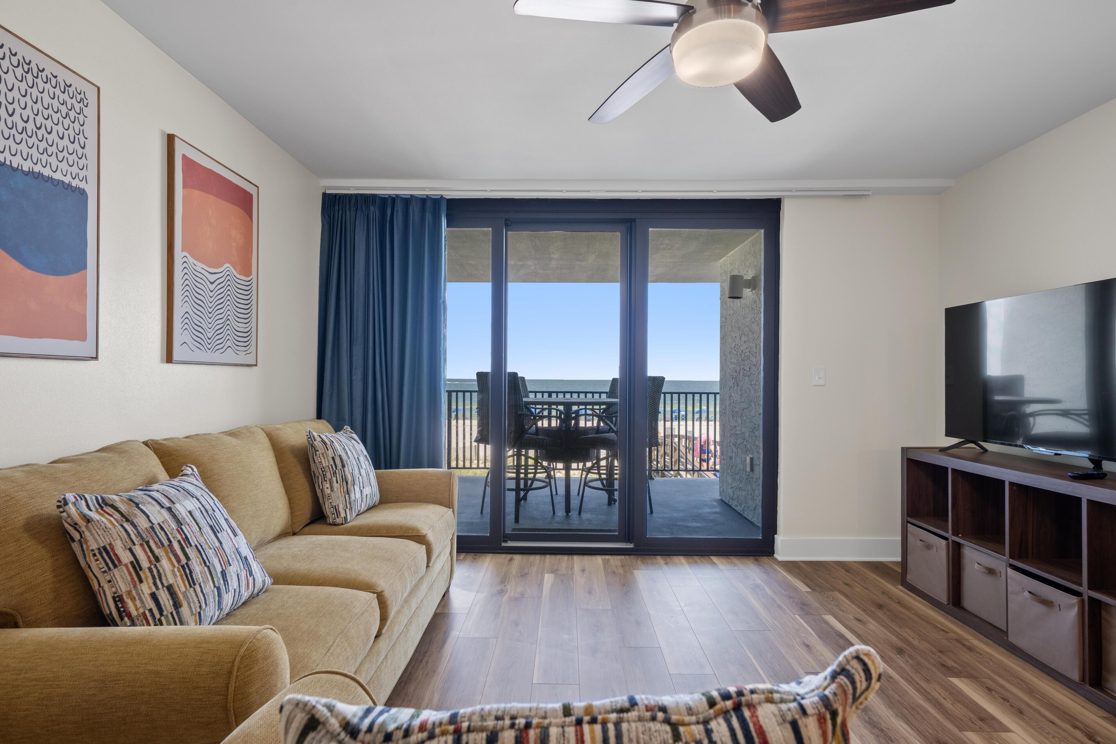 A living room with a sofa, a TV, a ceiling fan, and a balcony with a table and chairs.