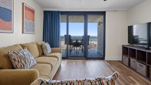 A living room with a sofa, a TV, a ceiling fan, and a balcony with a table and chairs. - Lei Lani 104 (Orange Beach)