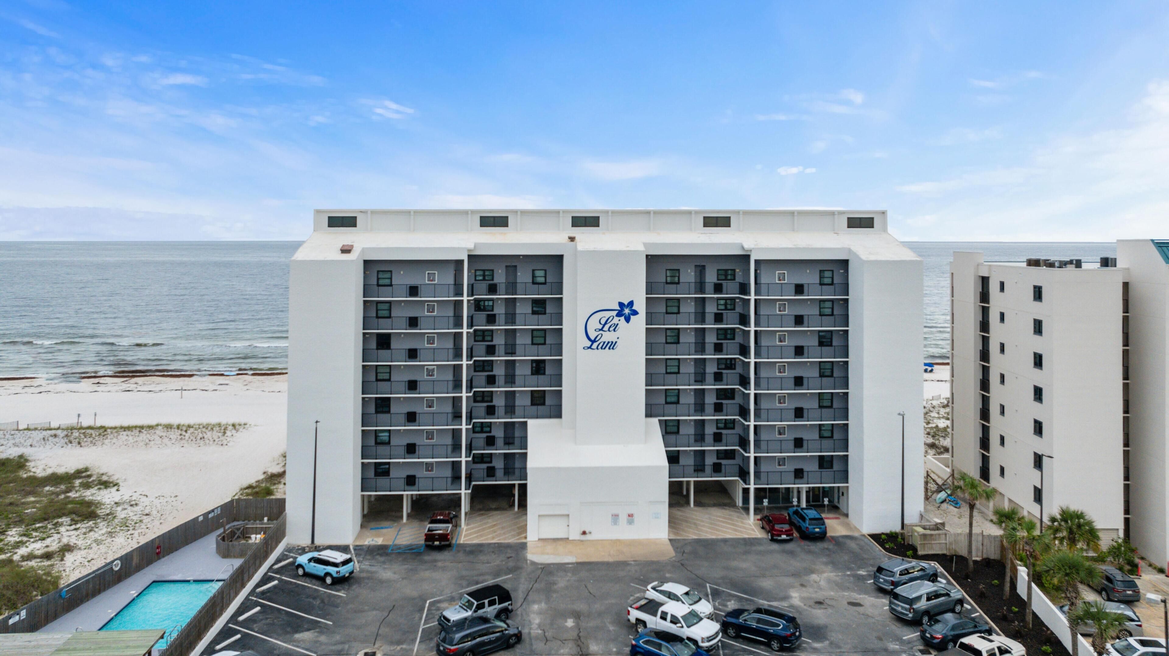 A multi-story hotel with a central entrance, parking lot, and a view of the beach and ocean.