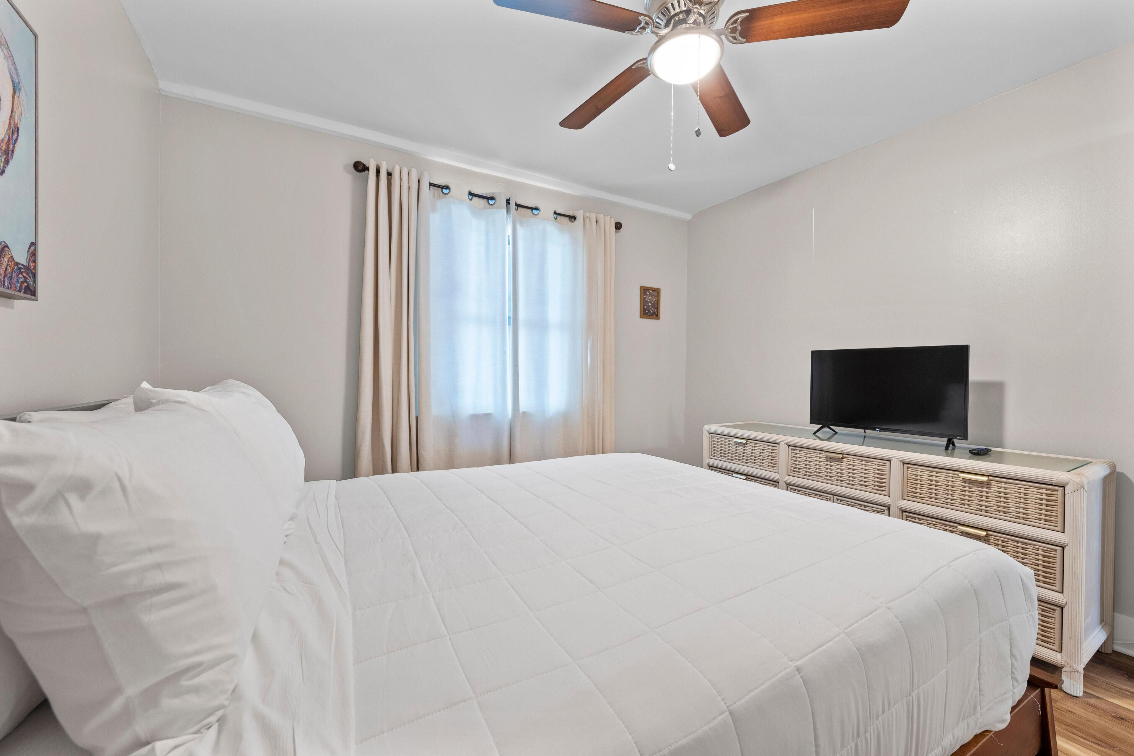 Une chambre à coucher avec un lit, un ventilateur de plafond, un téléviseur posé sur un meuble en bois et une fenêtre avec des rideaux.
