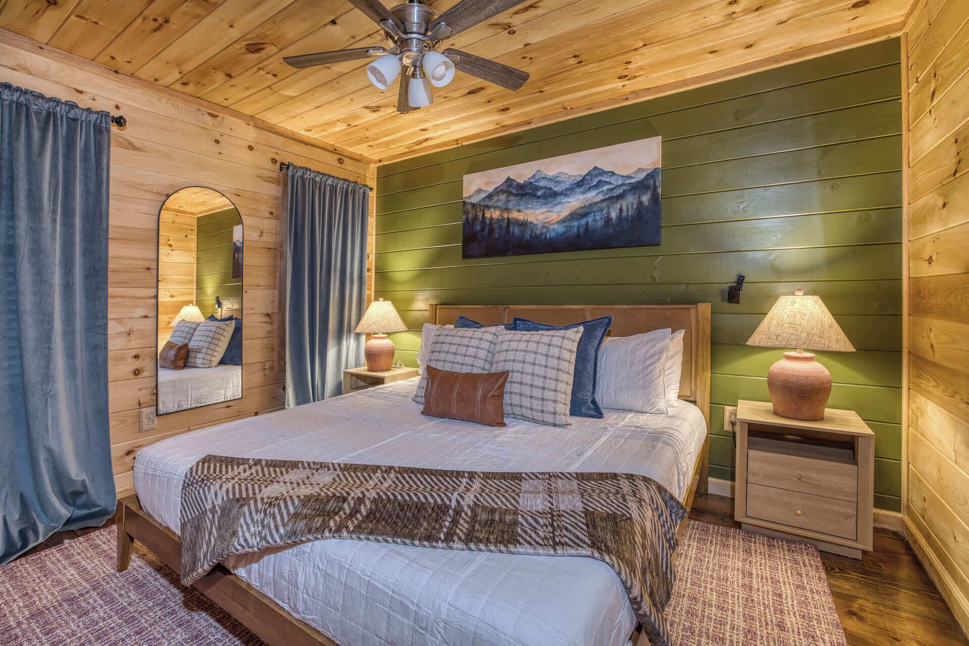 A wooden-paneled bedroom with a bed, two bedside tables with lamps, a ceiling fan, a large mirror, and a painting of mountains.
