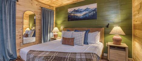 A wooden-paneled bedroom with a bed, two bedside tables with lamps, a ceiling fan, a large mirror, and a painting of mountains.