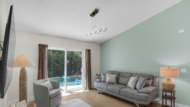 Una sala de estar con un sofá gris, una mesa redonda, una lámpara de pie y vistas a una piscina por un toldo corredero de cristal.