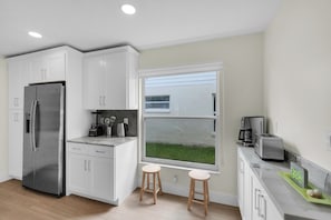 Eine moderne Küche mit weißen Schränken, einem Edelstahlkühlschrank, einer Kaffeemaschine und einem Fenster mit Blick auf eine Rasenfläche.