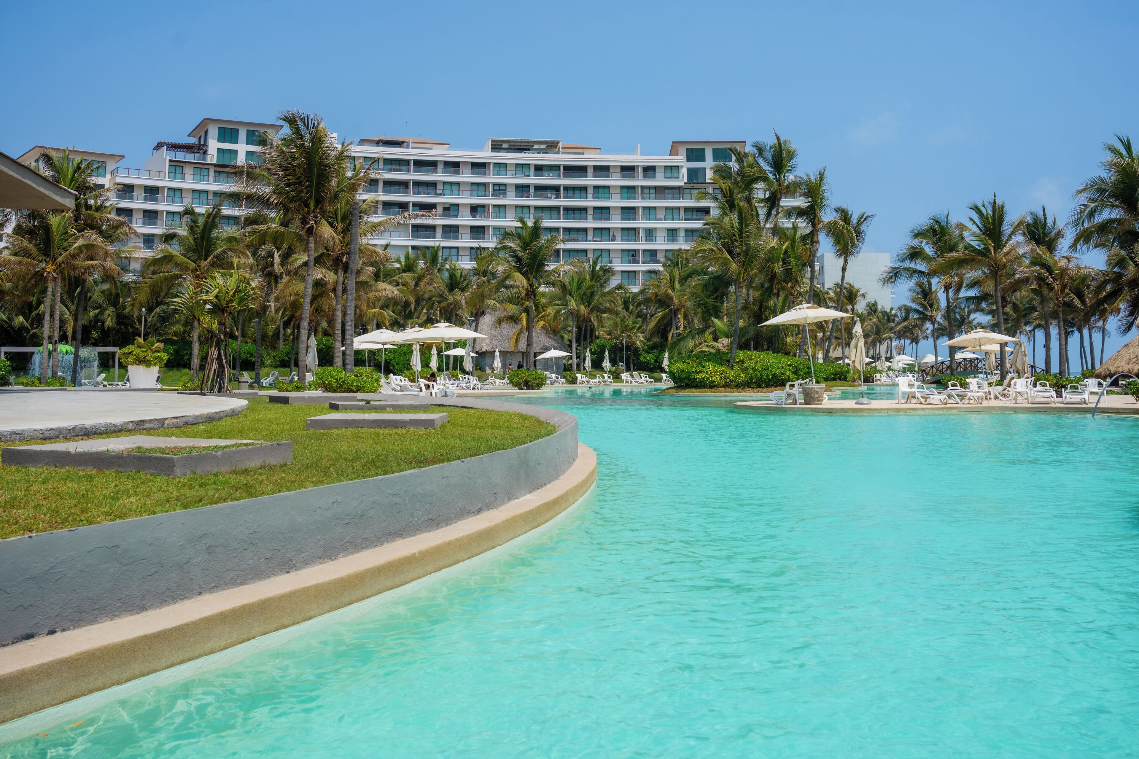 A resort pool with lounge chairs and umbrellas, surrounded by palm trees and a hotel building.