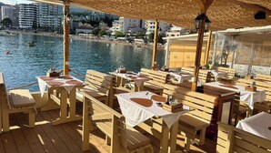 En restaurang vid havet med träbord och stolar, en överbyggd uteservering och utsikt över havet och byggnaderna i bakgrunden.