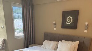 Ett hotellrum med en stor säng, en bänk, ett nattduksbord, en väggmonterad lampa och ett fönster med utsikt över byggnader.