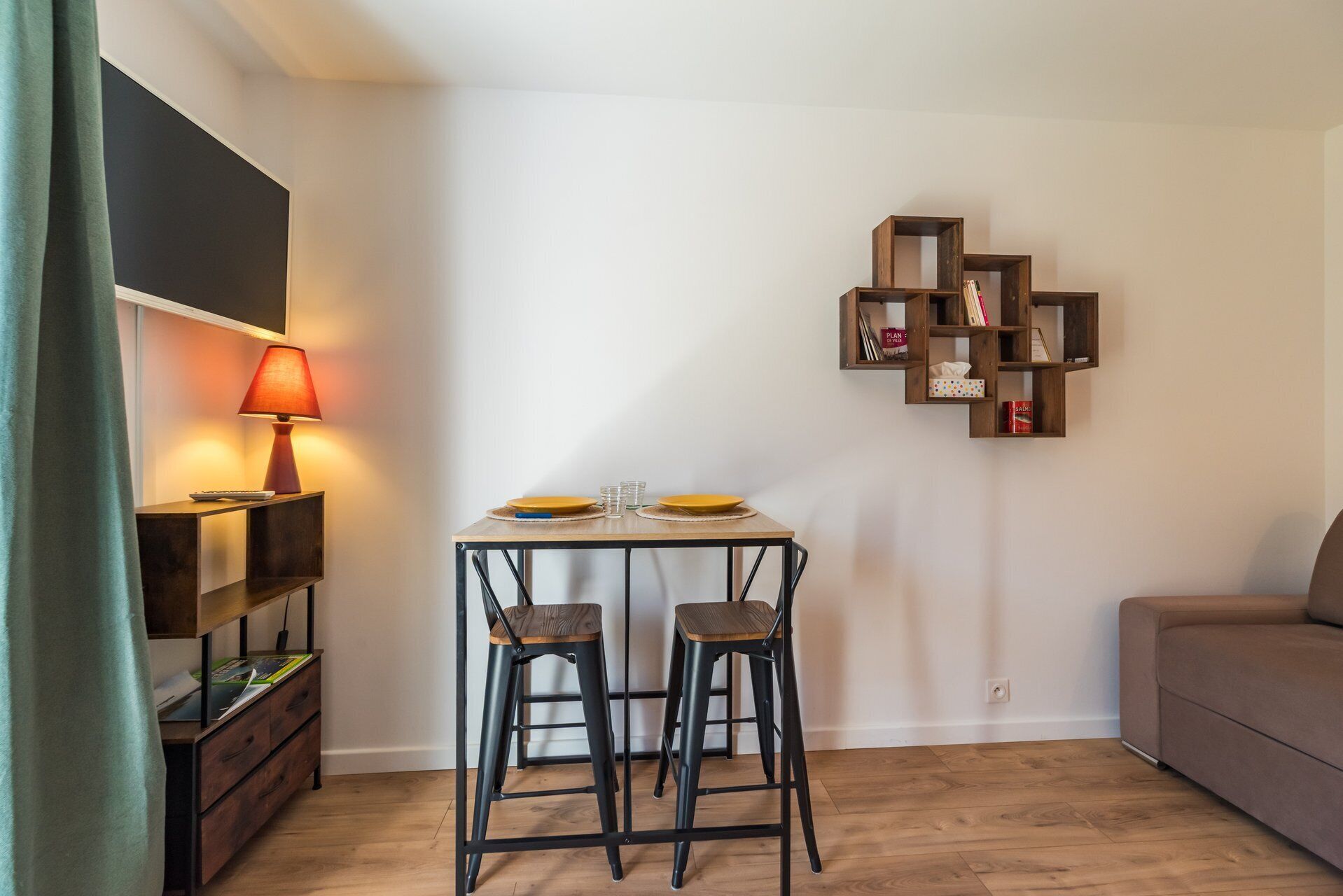 A small dining area with a table, two chairs, a lamp, and a wall-mounted shelf with books and items.