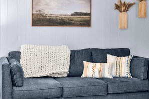 A grey sofa with a white knitted throw and patterned pillows, against a light blue wall with a framed landscape painting and two wooden vases with dried plants. - Jarrel Cottage Longer Stays Welcome! (Tyler)