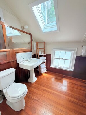 Un baño con tragaluz, piso de madera, inodoro blanco, lavamanos sobre pedestal y estante de madera con toallas.