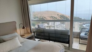 Ein Schlafzimmer mit Bett, Nachttisch, Lampe, Stuhl und einem Balkon mit Blick auf Stadt und Hügel.
