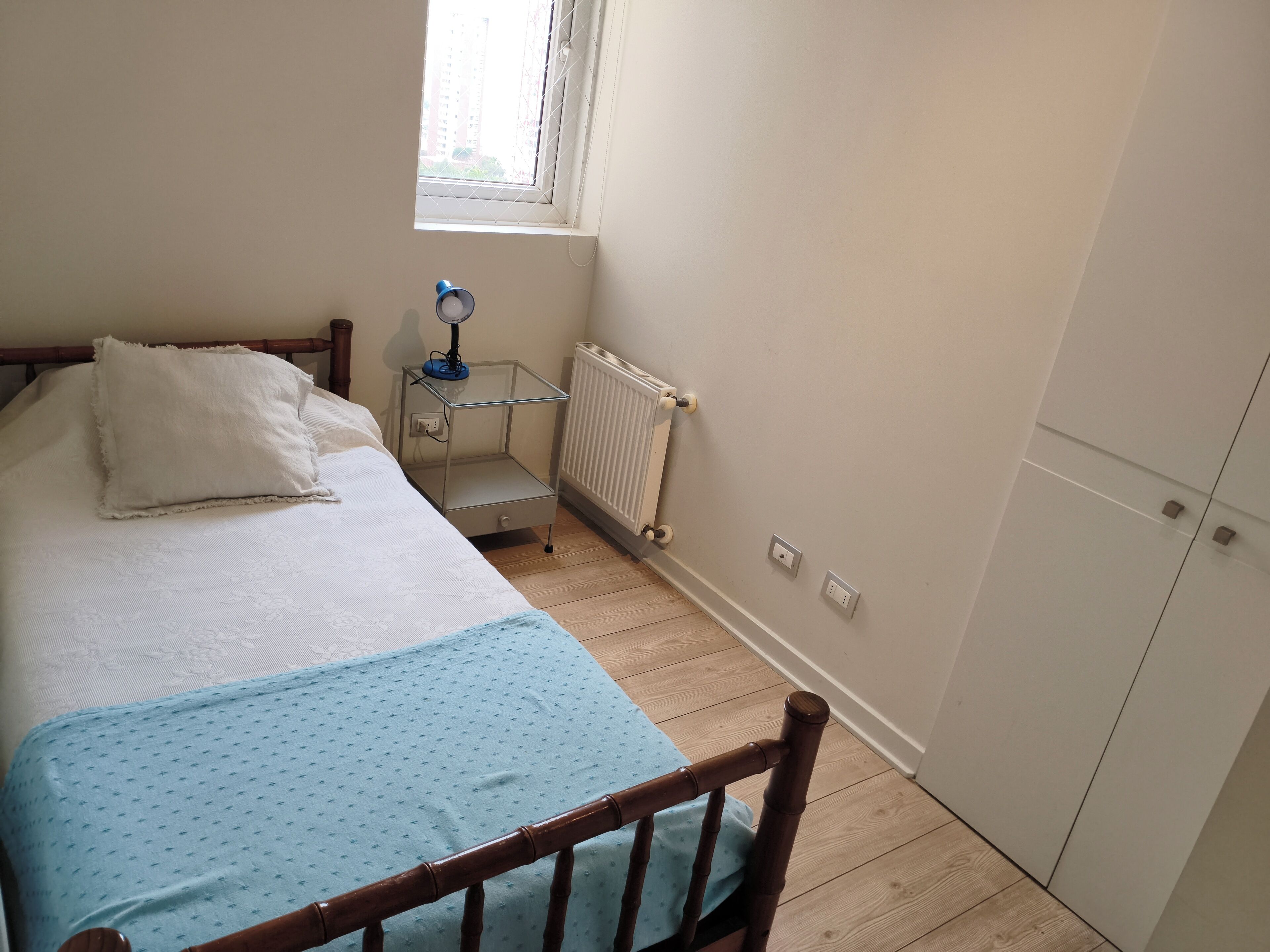 A bedroom with a bed, a nightstand with a lamp, a window, and a radiator.